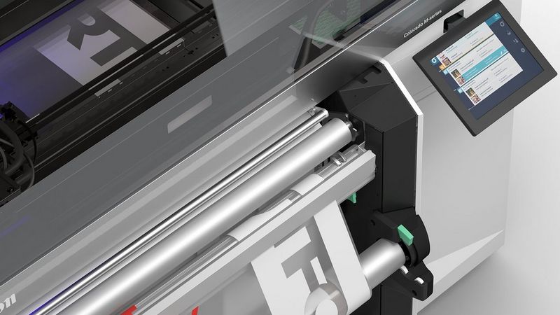 A close up of the printer rollers of a black and silver Canon Colorado M-series printer as it prints a white banner onto a clear substrate. There is a digital display to the right of the printed area.