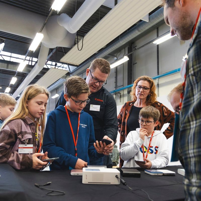 A group of children gather round an adult who shows them how to use a small Canon printer.