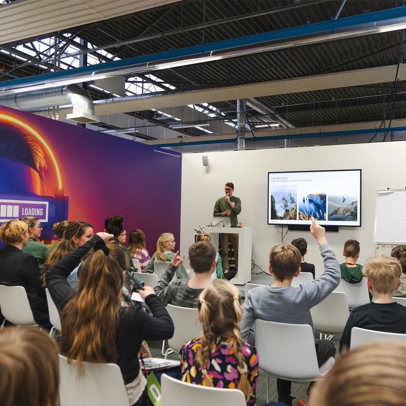 A man stands at a lectern, next to a screen and presents to an audience of young people whose backs are to the camera.