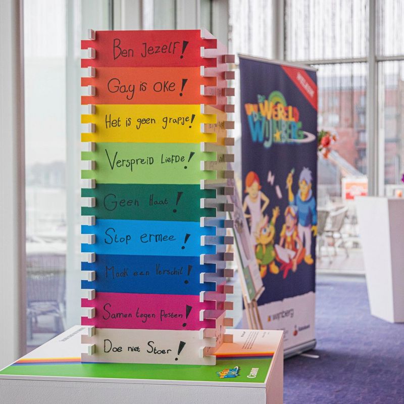 A three-dimensional printed piece, where four sides are connected by dovetail joints. It is striped in rainbow colours and each stripe has a different written slogan around inclusivity in Dutch, such as ‘ben jezelf!’ (be yourself!) and ‘gay is oke!’ (gay is okay!).