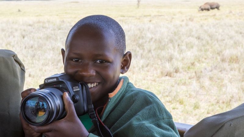 The head and shoulders of a smiling child sat in a vehicle. He wears a green fleece and holds a Canon camera. In the background is parched yellow grass and a pair of animals I n the distance. 
