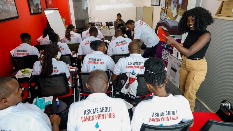 A classroom of students, photographed from the back of the class. They all wear white t-shorts with ‘Ask me about Canon Print Hub’ printed on the back. At the right of the shot is Monica Okech, who is smiling broadly as she speaks to a student.