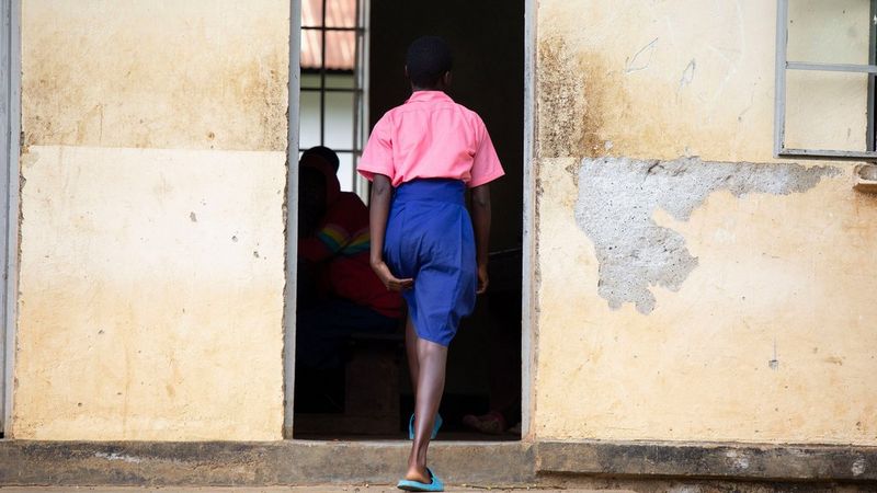 A woman, wearing a blue skirt and pink blouse, walks into a dilapidated looking building. She holds her skirt to her leg with her left hand.