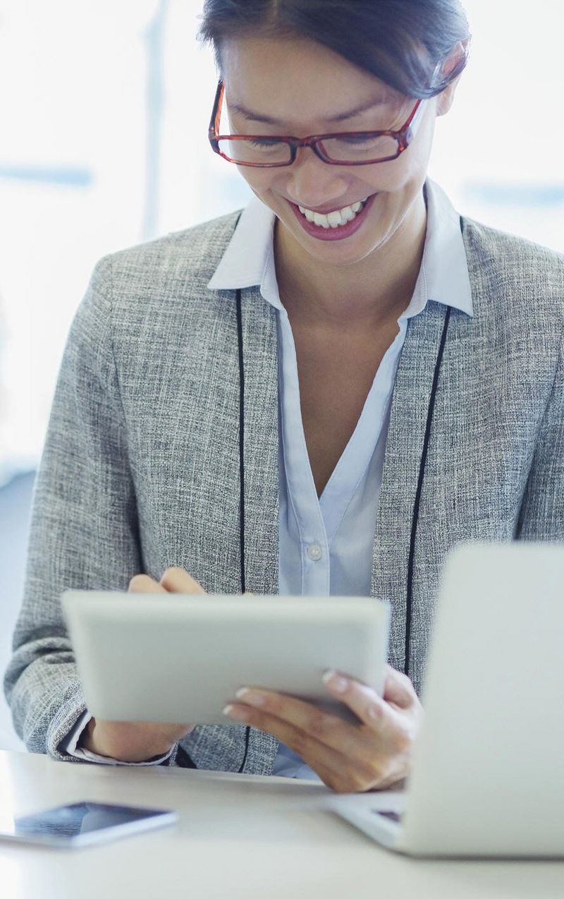 Woman with dark hair and glasses, wearing a light grey jacket and light blue shirt, smiles while using a tablet device.