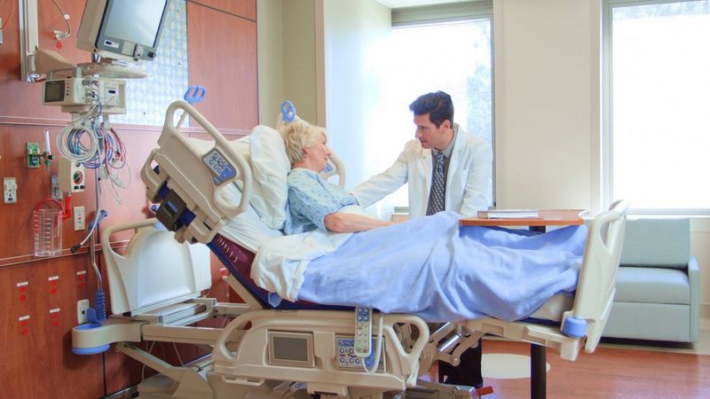 A doctor leans over a hospital bed to talk to an elderly woman patient.