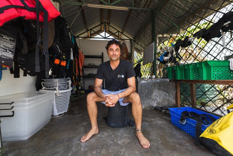 Le Dr Luca Saponari, pieds nus, en t-shirt et en short, est assis sur un seau retourné dans ce qui semble être un hangar à matériel. Il est entouré de boîtes en plastique et de matériel de plongée.