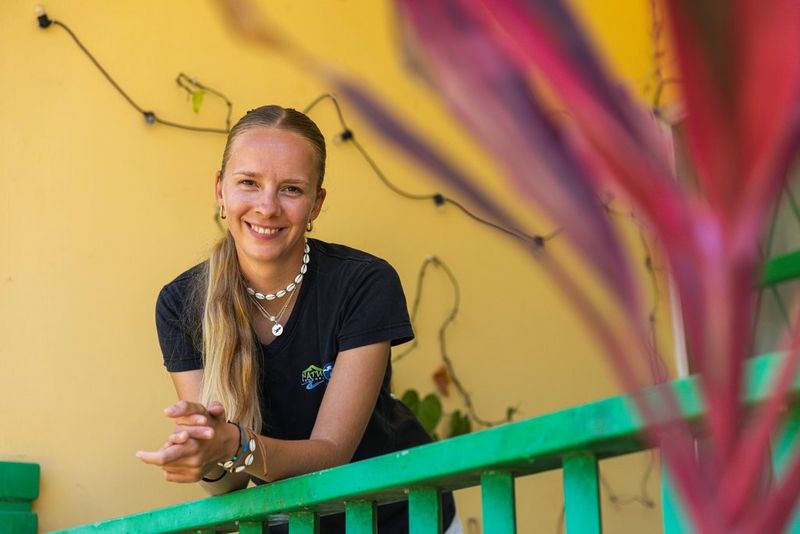 Vicky sourit et s'appuie sur une balustrade en bois vert devant un mur jaune vif. Elle porte des bijoux en coquillage. Au premier plan, à droite, se trouve une plante violette légèrement floue.