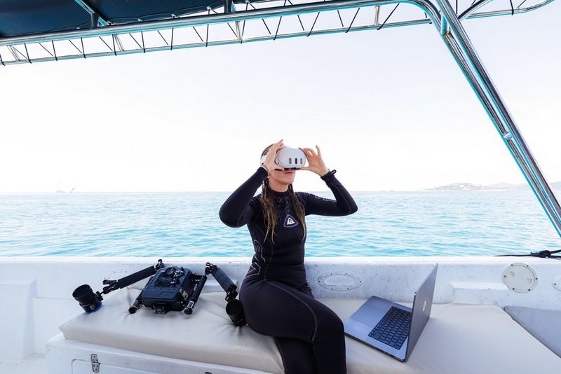 Мадлен Сен-Клер в гидрокостюме сидит на небольшом судне, надев VR-гарнитуру, а рядом с ней лежит MacBook Pro. Позади нее можно увидеть камеру Canon EOS R5 C с объективом RF 5.2mm F2.8L DUAL FISHEYE, которые закрыты в боксе для подводной съемки и установлены в крепление.