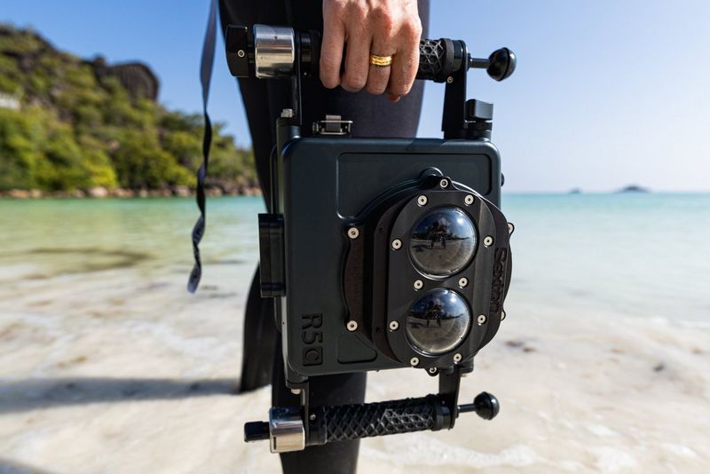 A woman in a wetsuit holds an underwater housing rig containing a Canon EOS R5 C camera with a RF 5.2mm F2.8L DUAL FISHEYE lens.