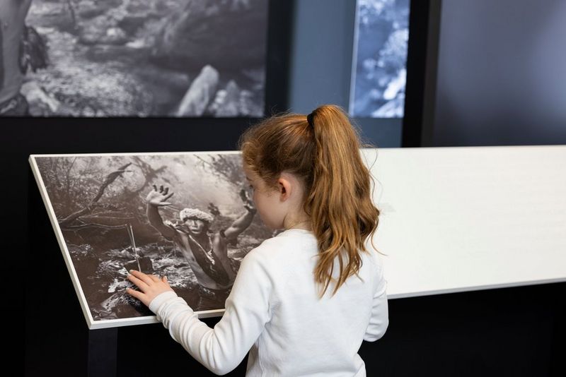 A child with a ponytail touches a black and white World Unseen exhibit image of an indigenous person in a forest.