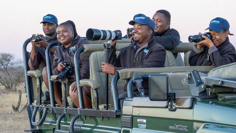 Wild Shots Outreach Programme Director, Rifumo Mathebula (centre) smiles broadly as he sits in a truck, holding a Canon camera with telephoto lens as he leads a game drive with his team of instructors, who are also holding Canon cameras.