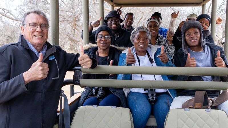 Wild Shots Outreach founder, Mike Kendrick (left), stands by a truck, giving the thumbs up. In the truck are a group of nine students, all holding Canon cameras, about to take their first ever game drive.