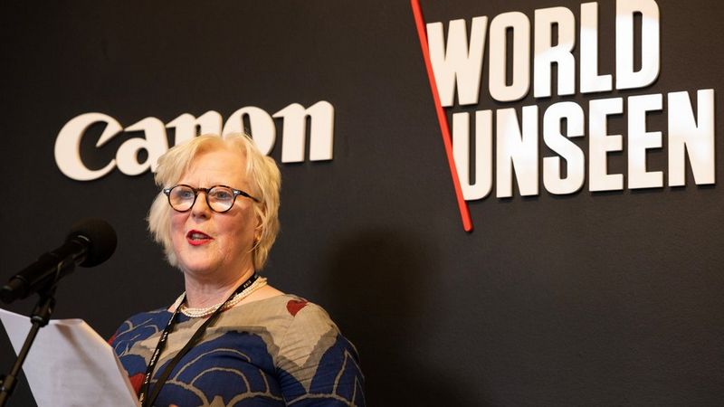 Anna Tylor, présidente du conseil d'administration du RNIB, se tient à gauche de l'image, devant un microphone et sur un fond noir sur lequel se trouvent des pancartes indiquant « Canon » et « World Unseen ». Elle a des cheveux blonds et porte des lunettes. Elle lit sur une feuille de papier.