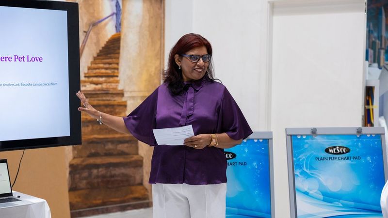 Eine Frau mit Brille und roten Haaren lächelt, während sie eine Präsentation hält. Sie trägt eine purpurfarbene Seidenbluse und eine weiße Hose. In einer Hand hält sie ein Blatt Papier, mit der anderen zeigt sie Richtung Bildschirm. 