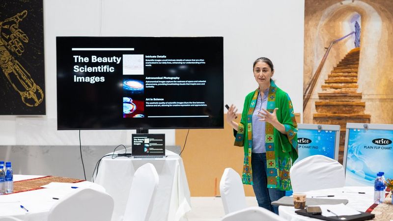 Eine Frau mit einer grün gemusterten Jacke über einer hellen Bluse und Jeans hält eine Präsentation. Sie steht neben einem großen Bildschirm, auf dem der Titel „The Beauty Scientific Images“ zu sehen ist.