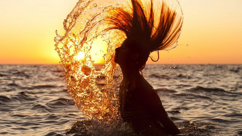 A woman in the sea at sunset flicks back her hair, creating an arc of water spray.