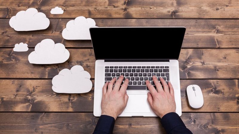 Cloud computing technology concept with cut outs of white fluffy clouds sat to the left of the laptop. A pair of hands type on the keyboard, which is sat on a dark brown wooden table. 