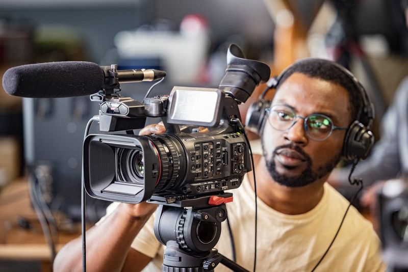A man wearing headphones filming on a Canon XF605 camcorder with a microphone attached.