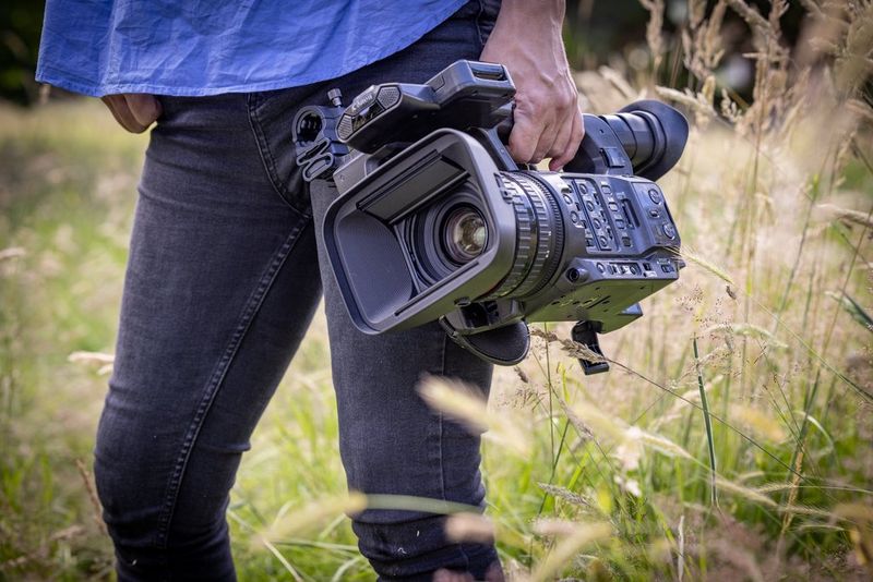 A close-up of a person carrying a Canon XF605 camcorder as they walk through a field.