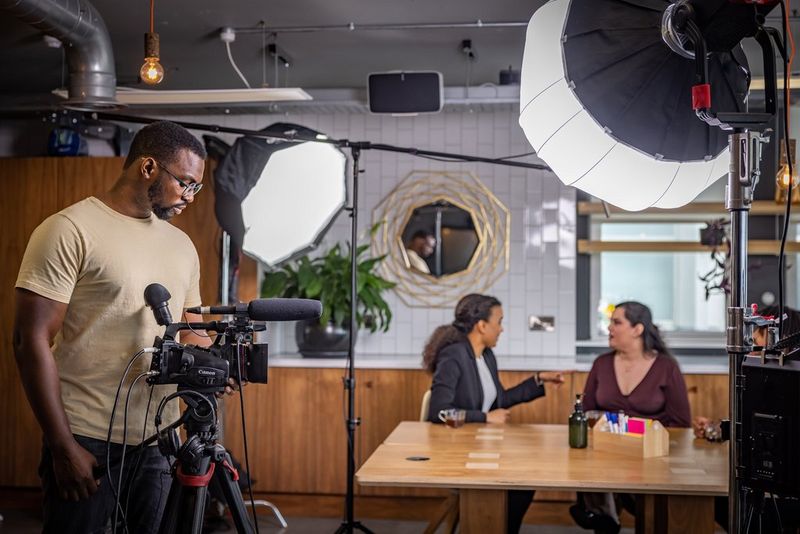 Мужчина в окружении оборудования для вещания снимает двух людей, которые обсуждают что-то, сидя за столом.