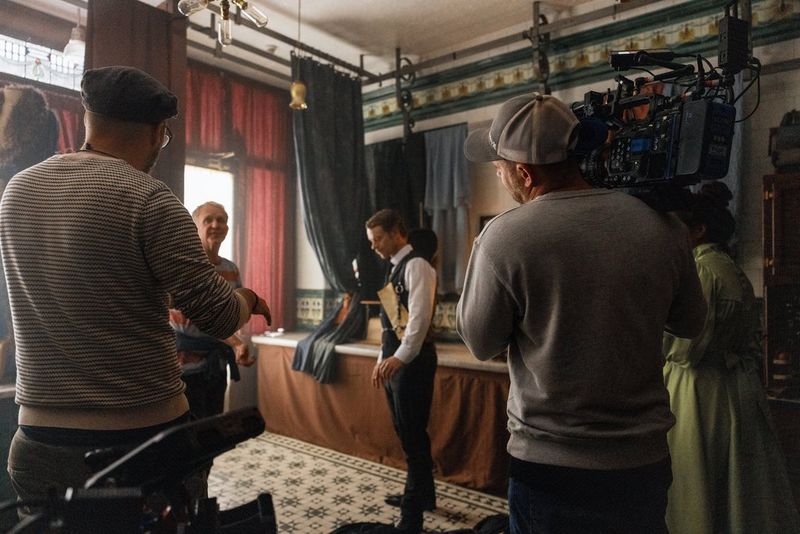 DoP Fabian Wagner films inside an Edwardian dressmaking shop with a Canon EOS C500 Mark II on his shoulder. A man wearing smart black clothes stands just in front of the camera.