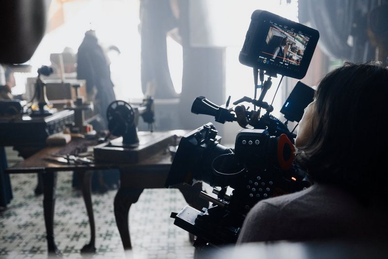 A large cinema camera is set up in the corner of an old-fashioned dressmaking shop.