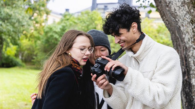 Un jeune homme sourit en montrant une photo sur l’écran de son appareil photo Canon à deux jeunes femmes dans un parc.