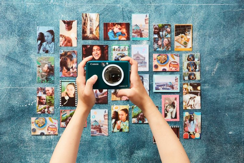 Une personne avec un appareil Canon Zoemini S2 photographie un mur recouvert de nombreuses photographies miniatures de deux jeunes qui s'amusent.