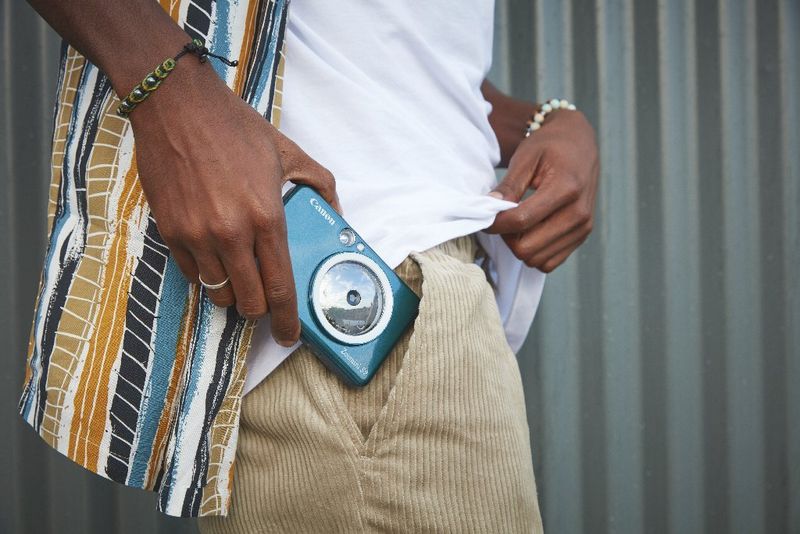 Un homme vêtu d'un pantalon beige et d'une chemise colorée glisse un appareil Canon Zoemini S2 dans sa poche.