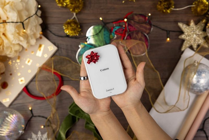 Человек держит в руках компактный принтер Canon Zoemini 2 с маленьким красным бантом. На заднем плане можно увидеть праздничные украшения и гирлянды.