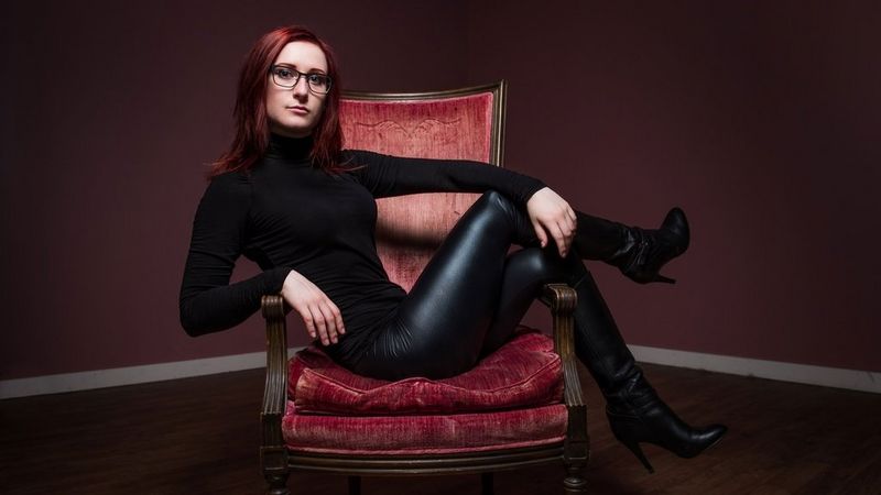 A bespectacled Zoë Rose sits crossways on a high-backed chair, upholstered with red velvet. Her back leans against one arm and her legs rest over the other. She has long red hair and wears a red polo-neck, black shiny leggings and high heeled ankle boots.