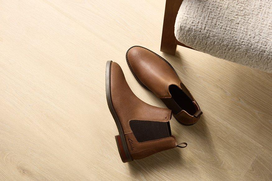 A pair of Clarks’ Jaxen Easy boots in Cola Suede (brown) on a tan hardwood floor at the foot of an upholstered task chair