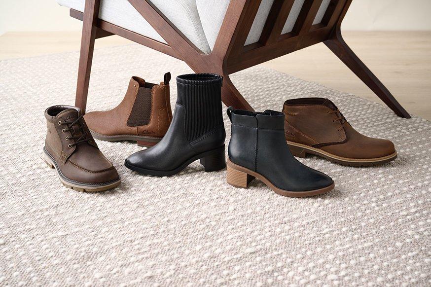 An array of black and brown men and women’s boots at the foot of an upholstered accent chair on a cream-colored rug