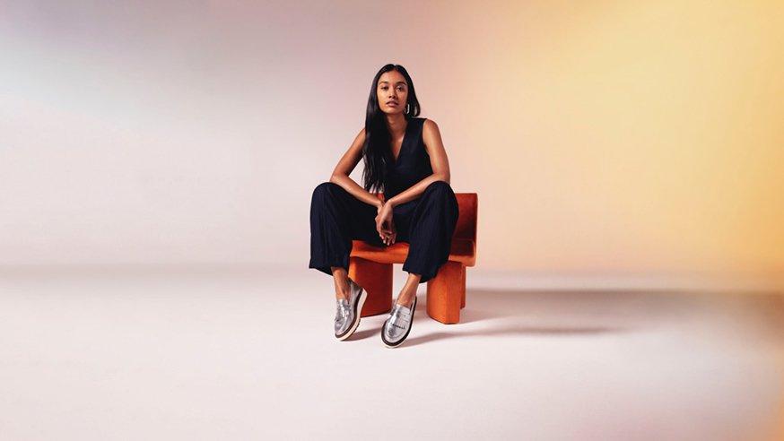 Young woman sitting on an orange chair wearing a wide-leg jumpsuit and Clarks’ silver leather Orianna loafers