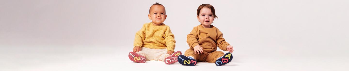 Baby boy and girl sitting next to each other with their legs stretched out showing the shoe sole in pink and blue with letters and numbers