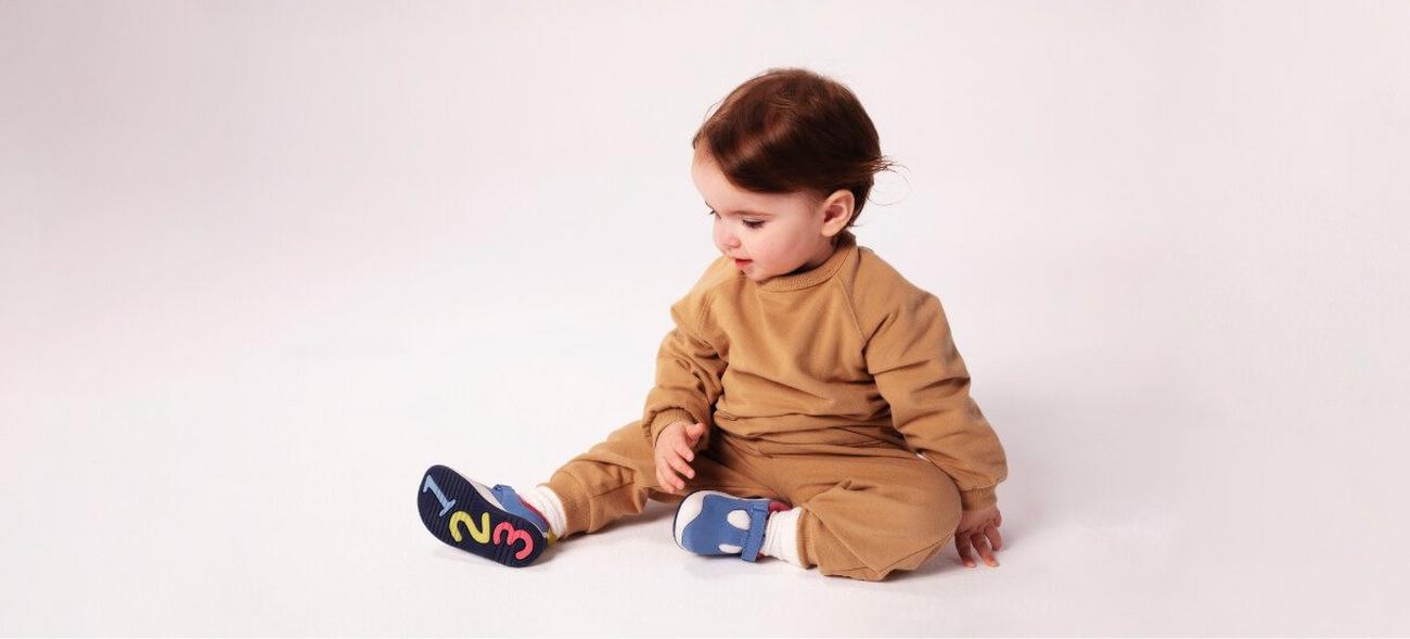 Baby girl sitting with her legs stretched out looking at her shoes that have letters on the sole