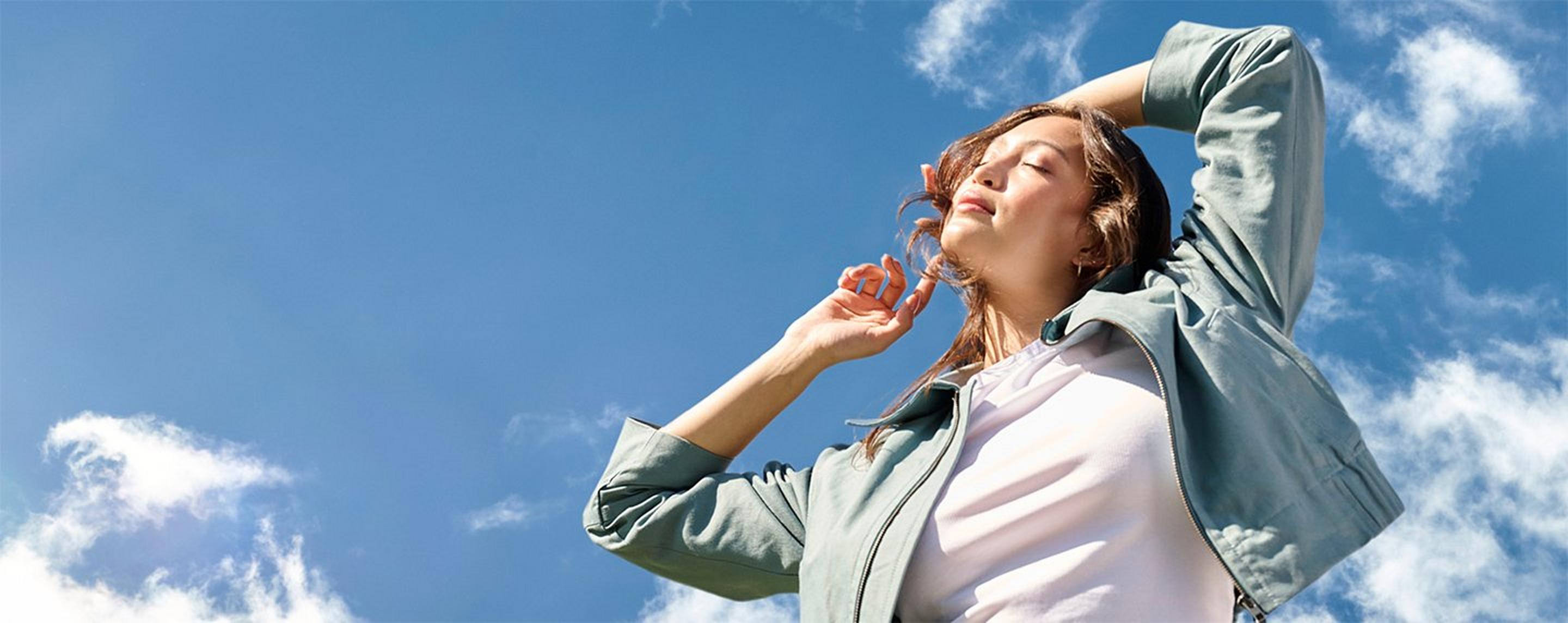 A woman holding her hands in the air looking up at the sun on a blue sky with clouds.