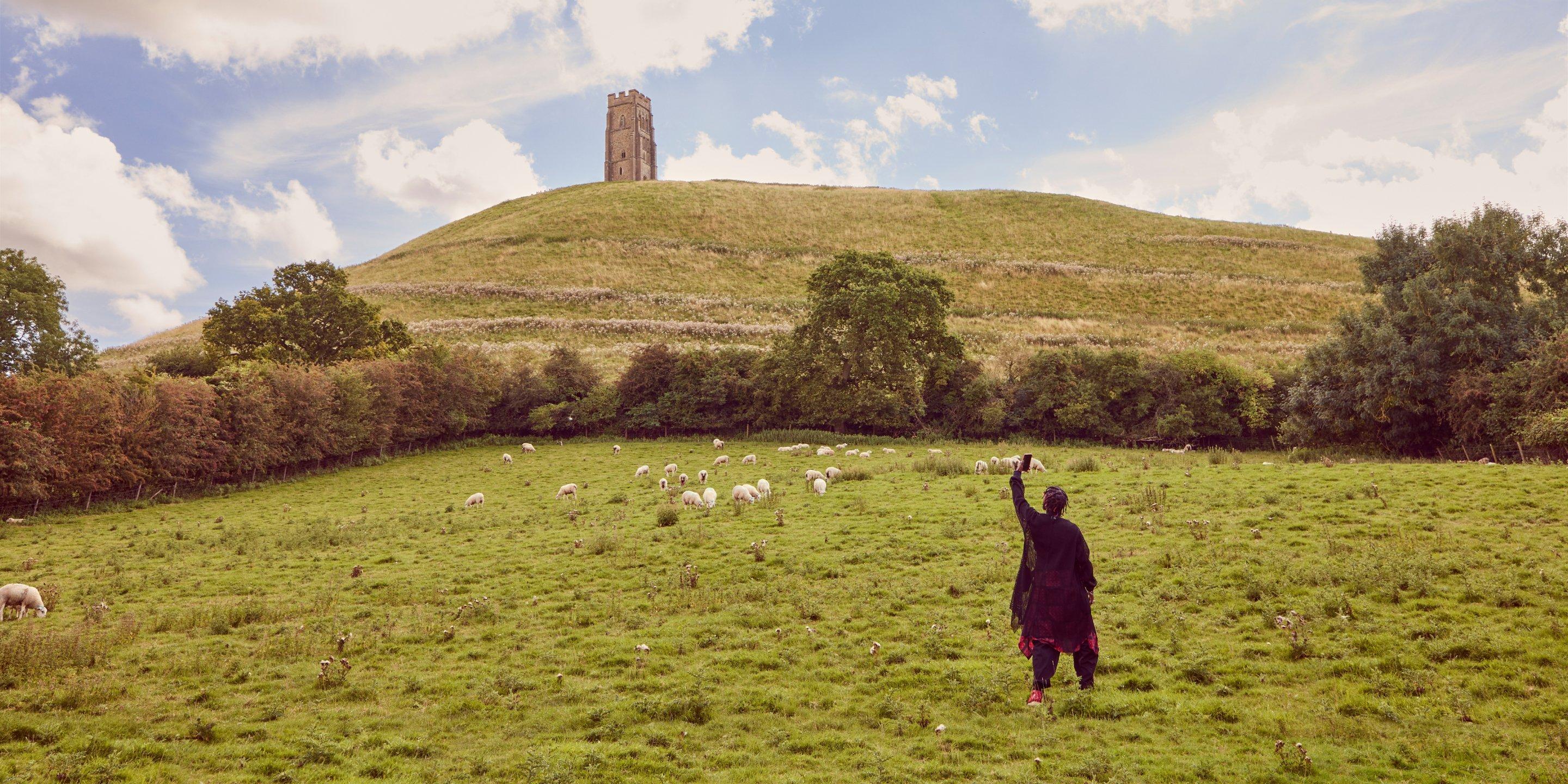 Popcaan debout dans un champ avec des moutons en arrière-plan prenant une photo de Glastonbury Tor