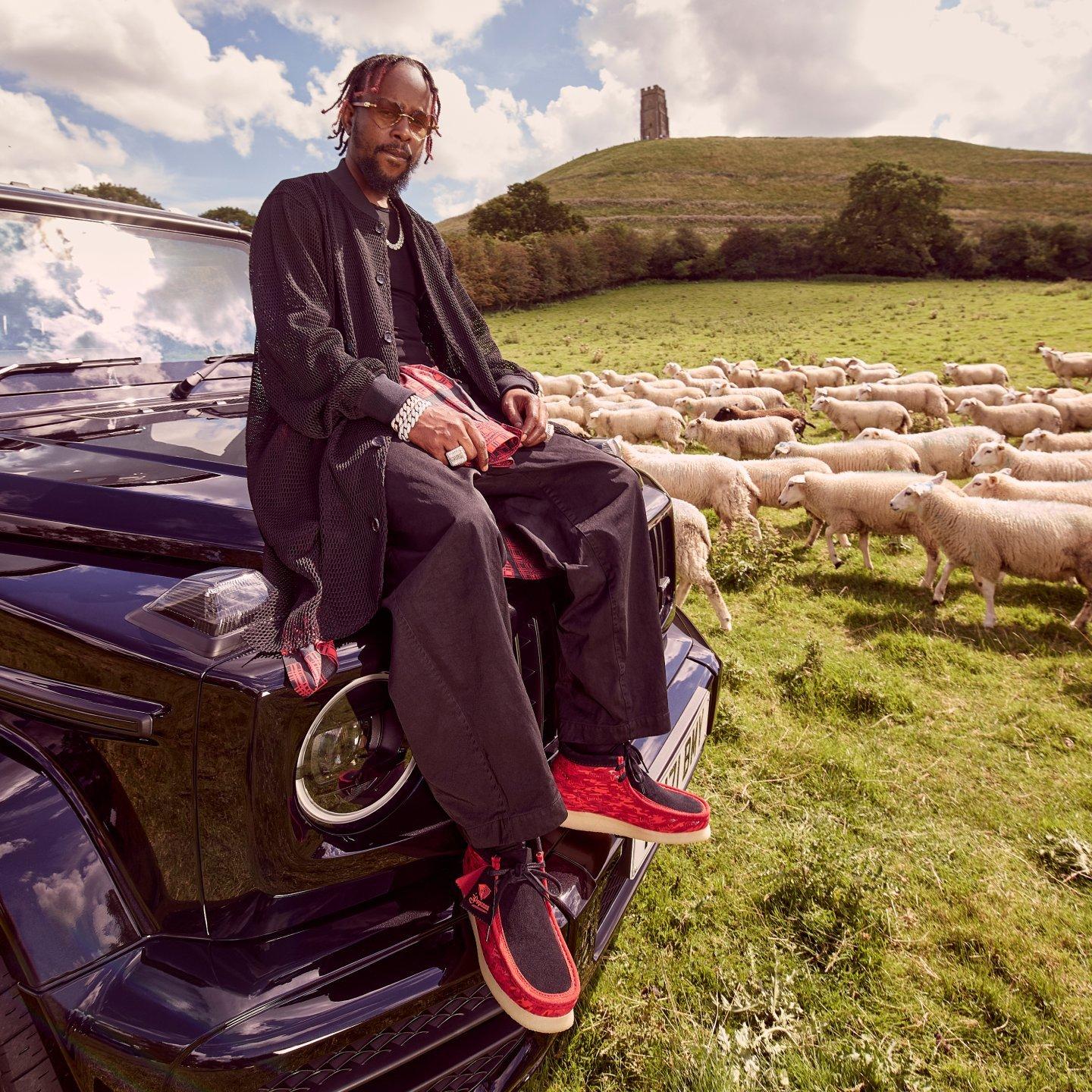 Popcaan assis sur un chapeau Jeep regardant la caméra avec la Glastonbury Tor en arrière-plan.