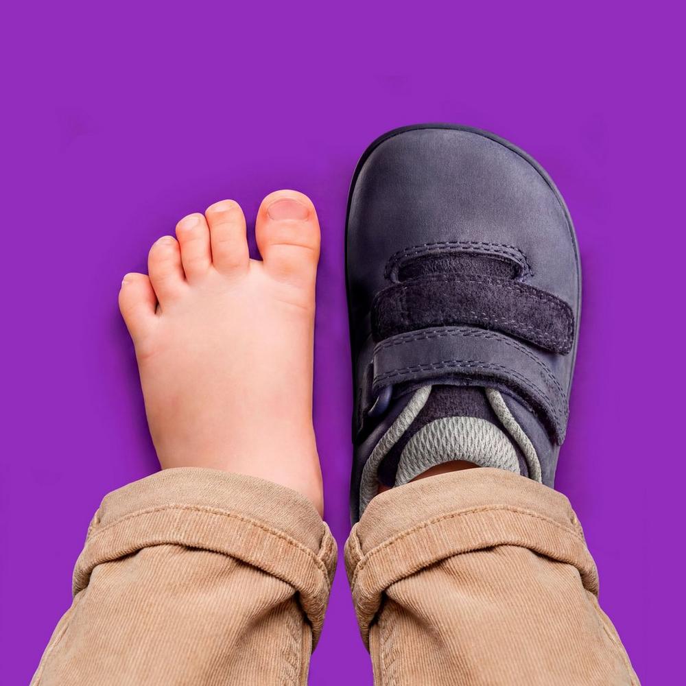 A child's bare foot beside a child wearing a black Clarks shoe