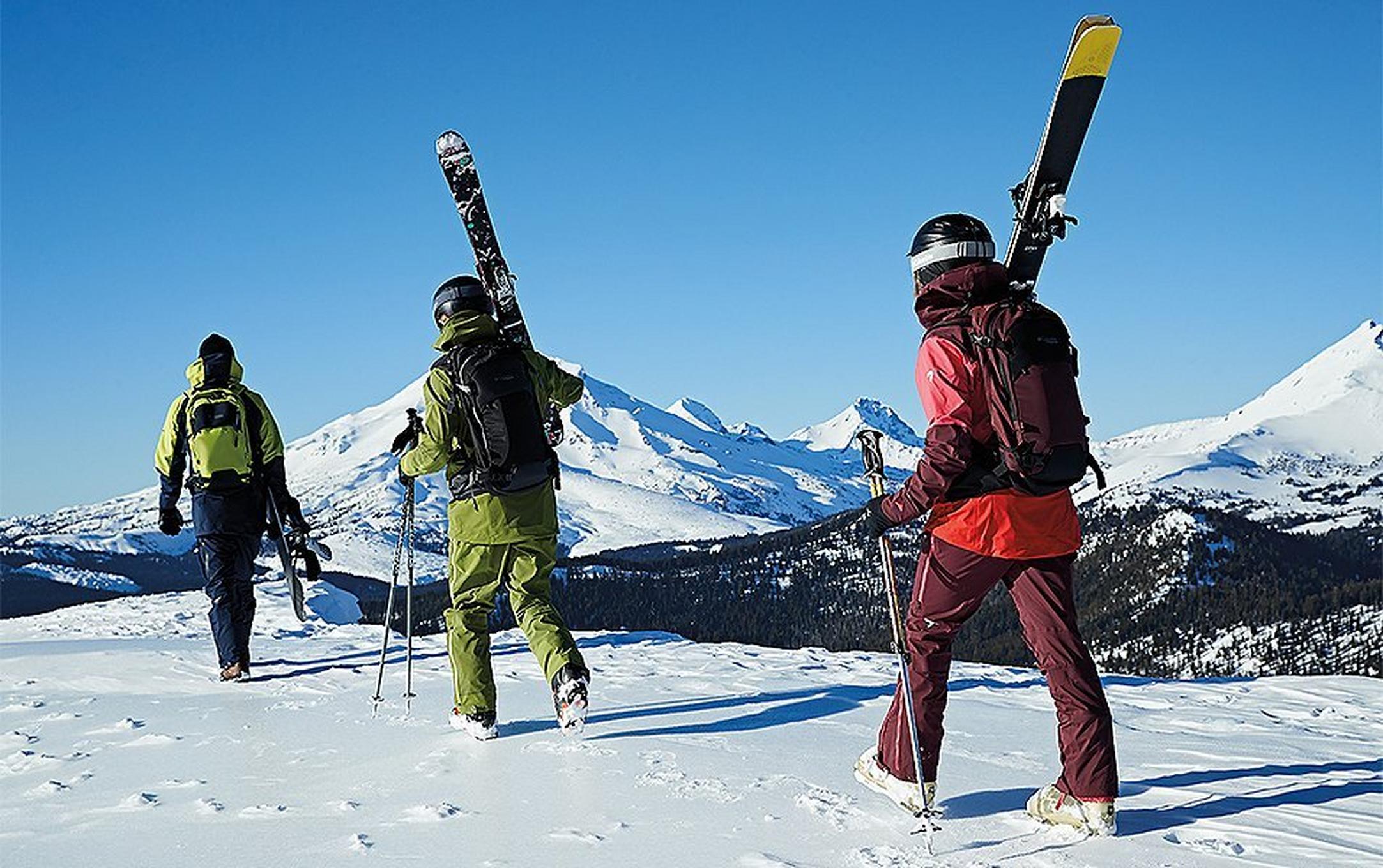 Three skiers walk up the mountain in a single file line on a gorgeous sunny day.