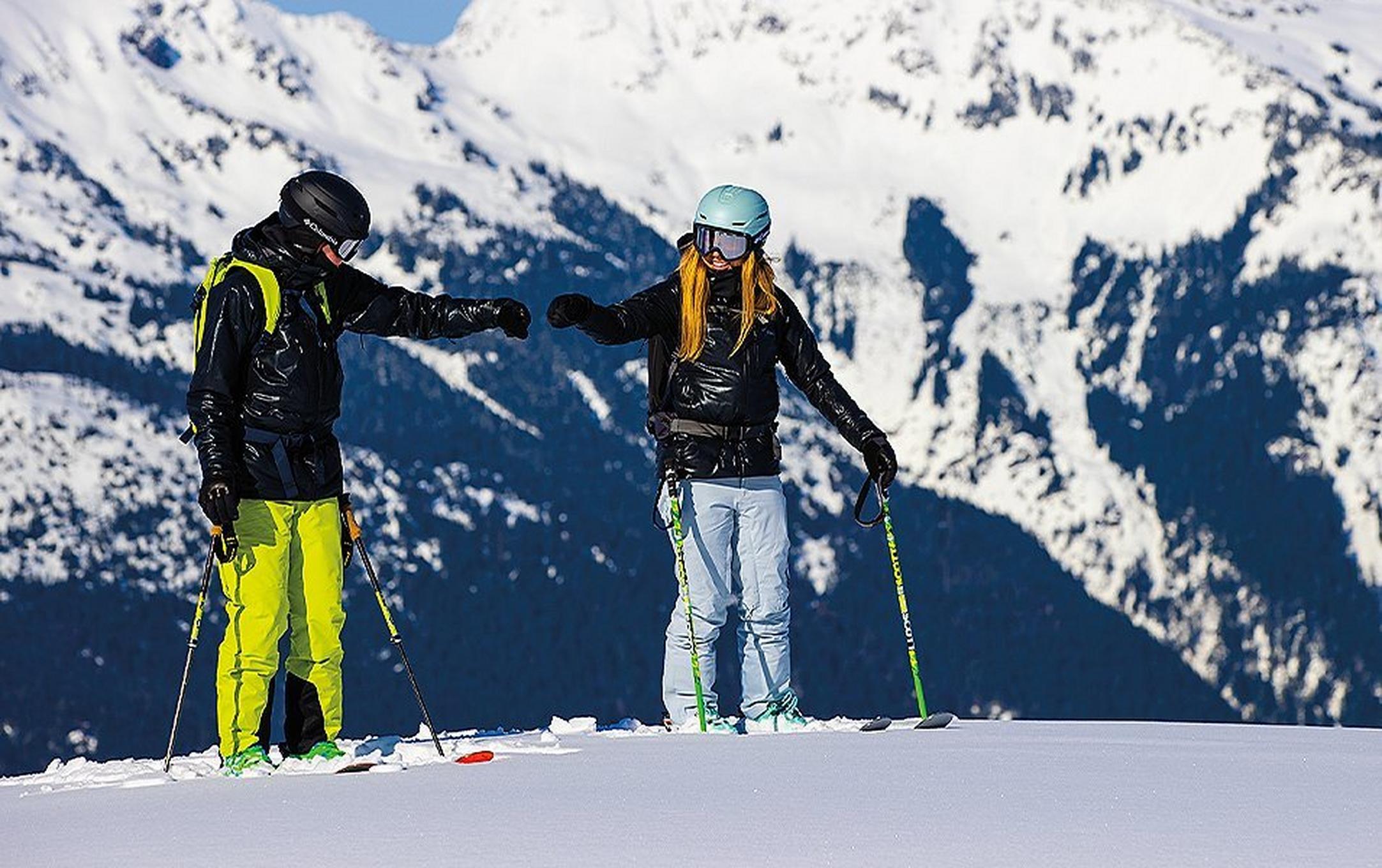 Two skiers reach out and bump fists as they smile on top of a ski run.