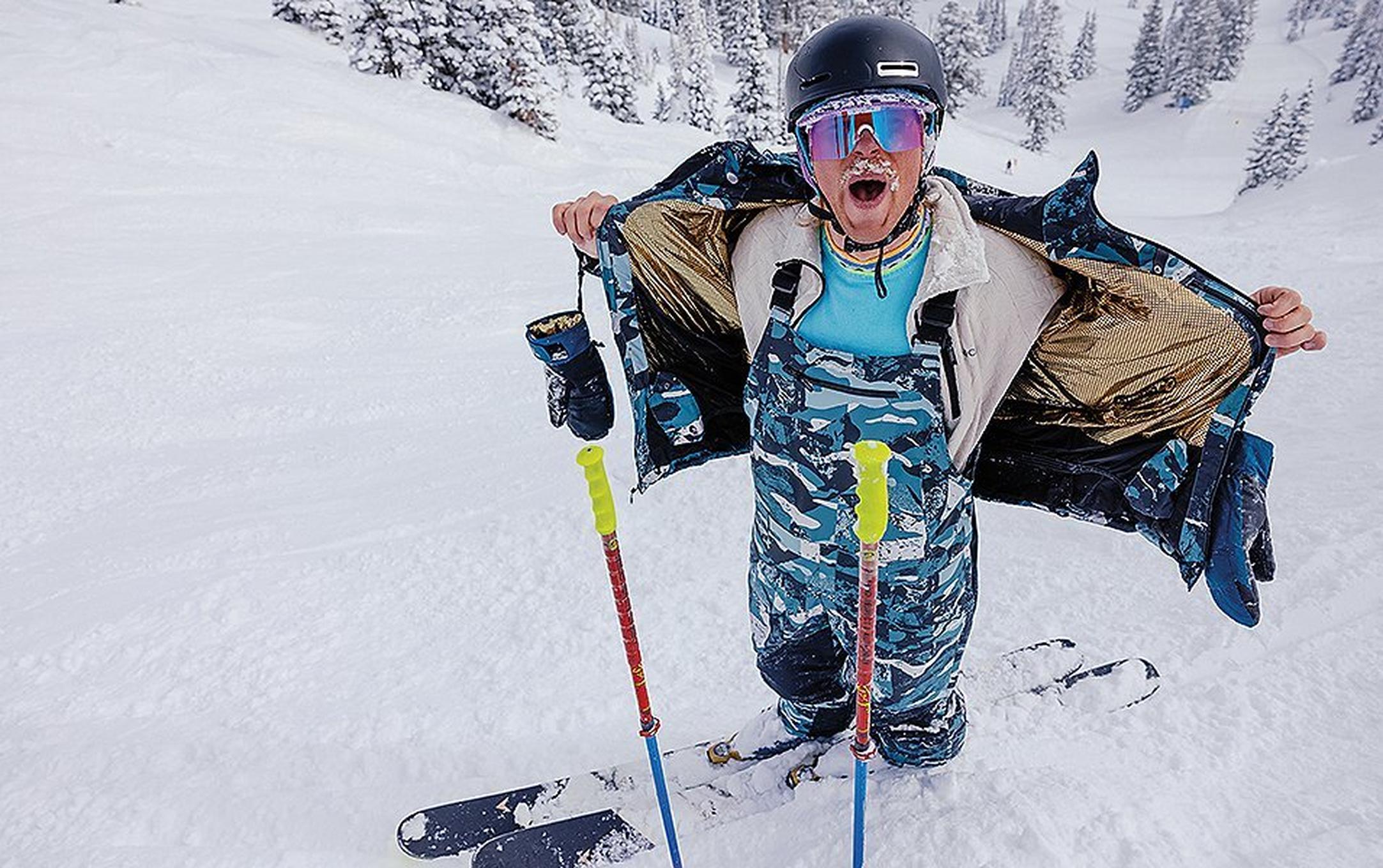 A skier with snow caked in his mustache removes his ski coat as he stands on a powdery ski run.