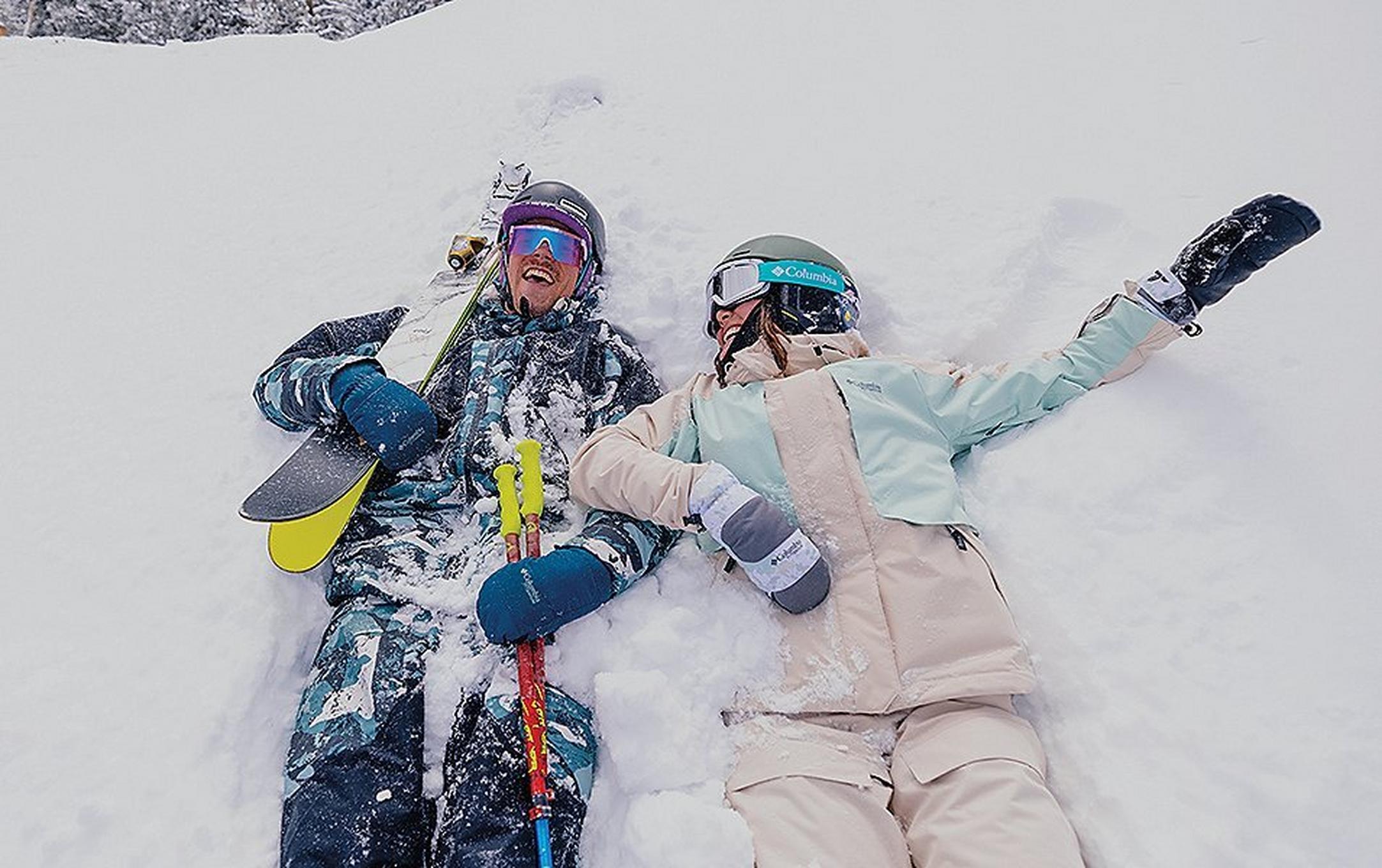 Two skiers lie on their backs in a field of snow as hey smile and laugh together.