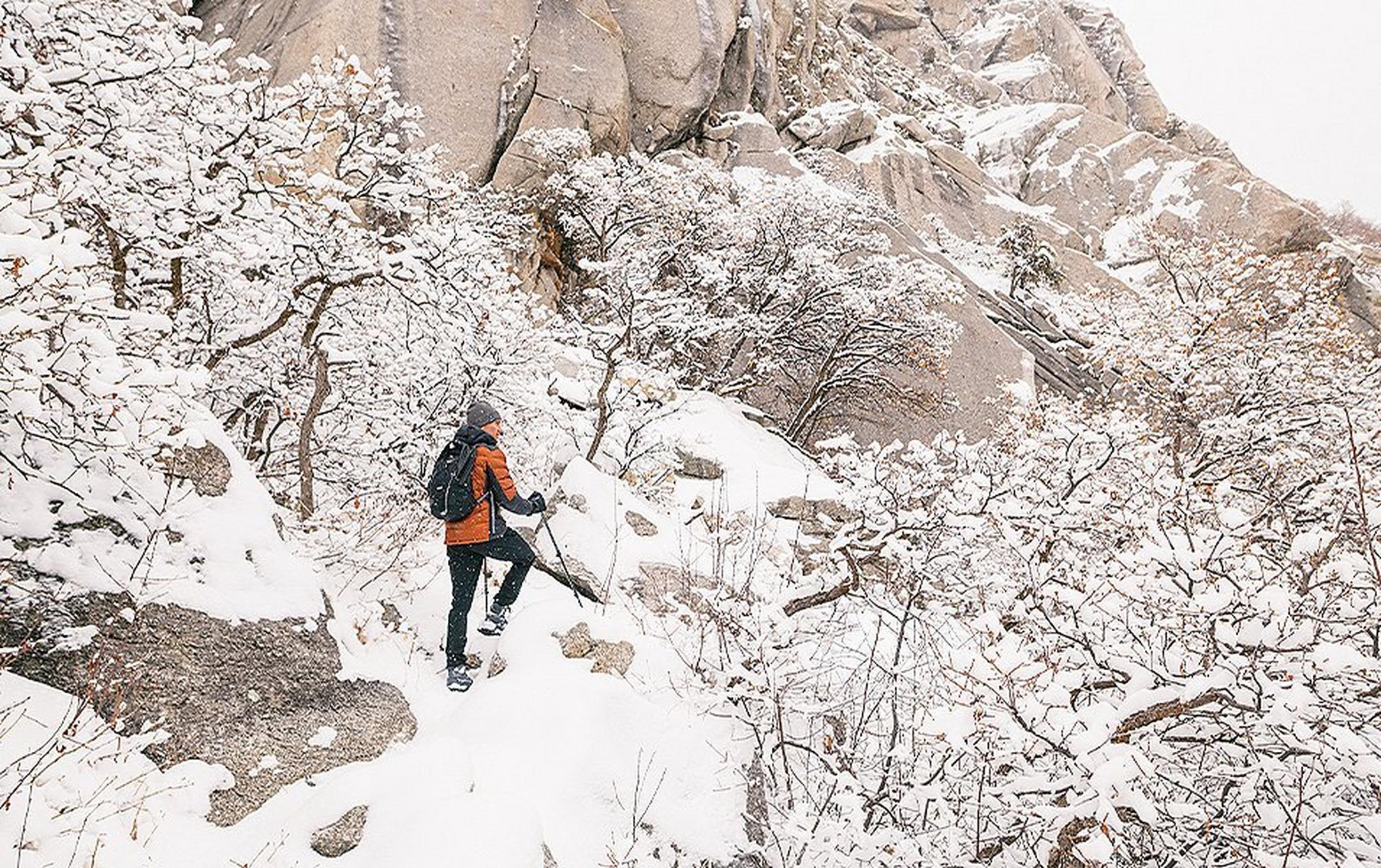 A bundled-up man in an orange puffy jacket and black snow pants stands with trekking poles on a snowy cliff watching the scenery.