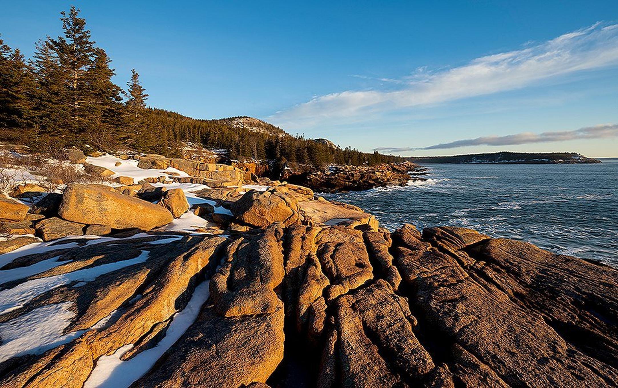 Golden rocks covered with pockets of snow sit next to the deep blue ocean as the morning light hits them in Acadia National Park.