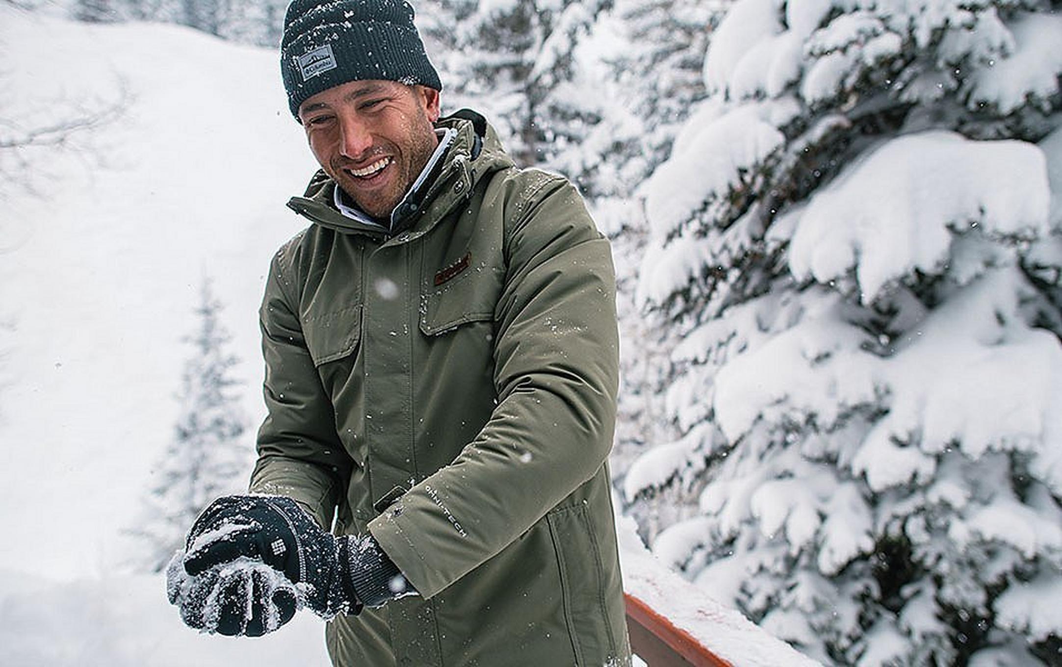 A man in an olive green jacket grins widely as he makes a snowball in his hands which are donning black winter gloves.