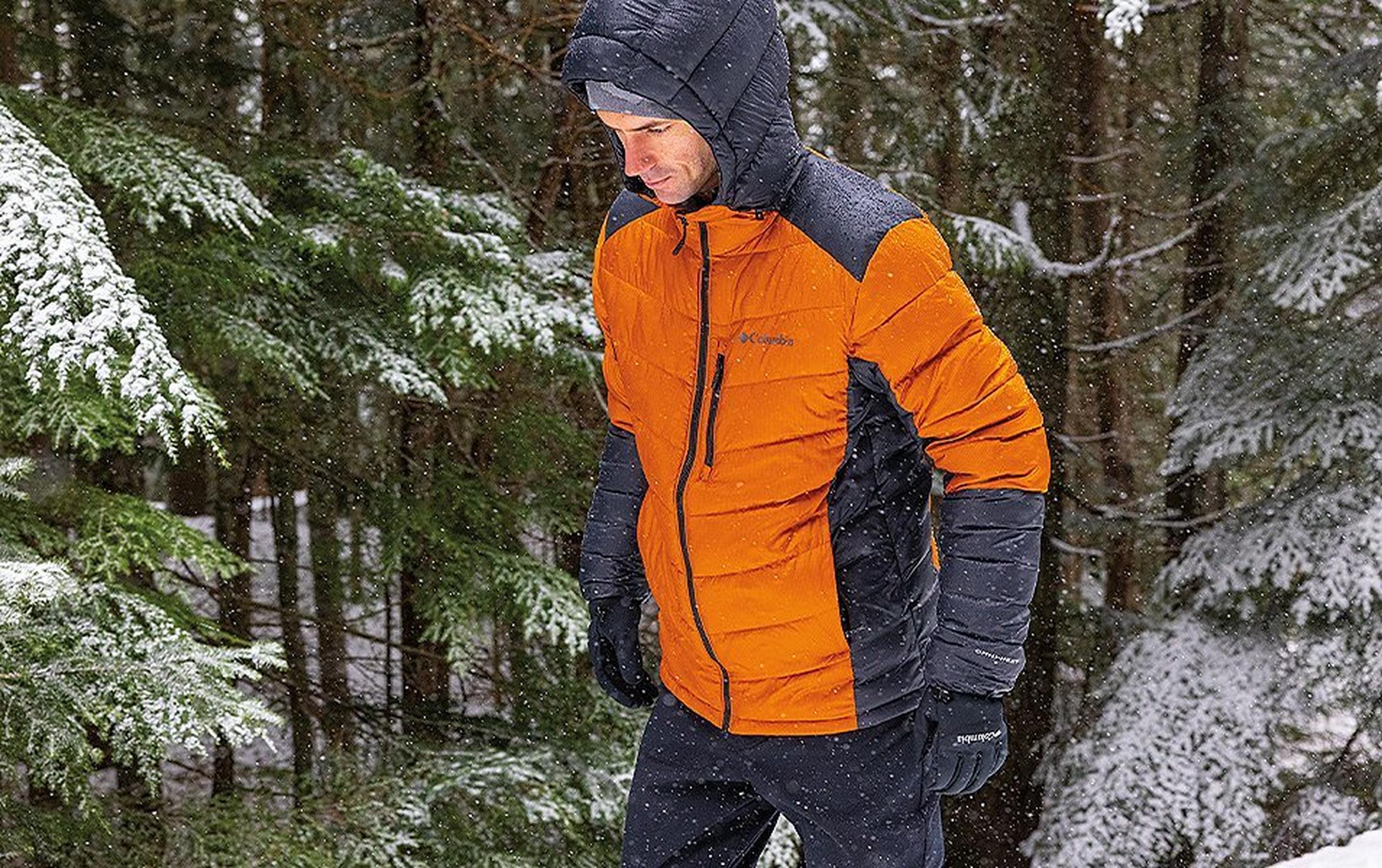 A man wearing an orange Columbia Sportswear puffy coat walks through the woods.
