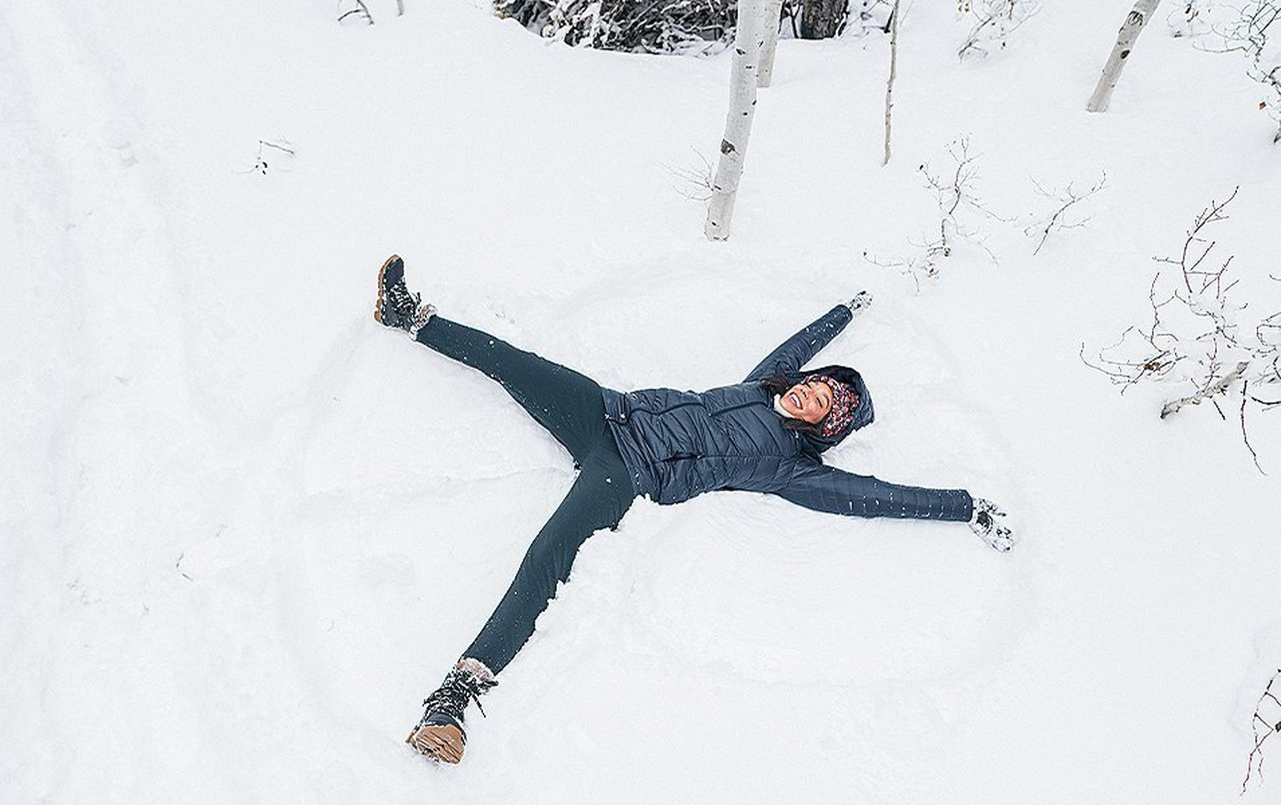 A woman wearing a puffer coat smiles as she makes snow angels on the ground in a winter setting.