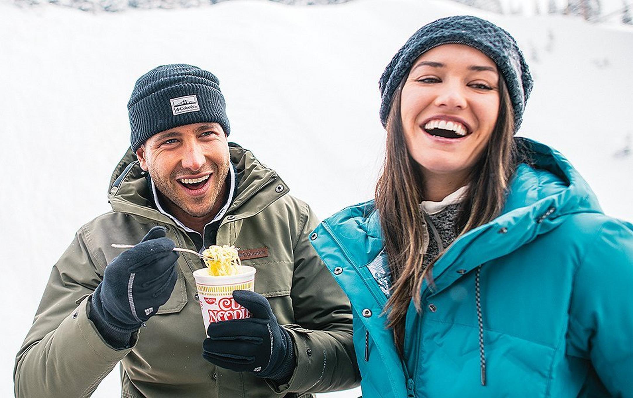 A man in an olive-colored jacket stands with a woman in a blue puffy coat as he eats a Cup of Noodles and they laugh together.